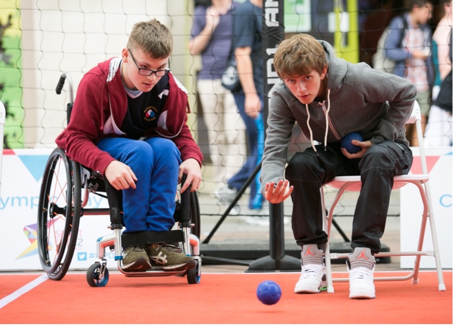 Image of two boys playing boccia. One boy is a wheelchair user and the other is seated on a chair, with his arm outstretched having just bowled the ball. 